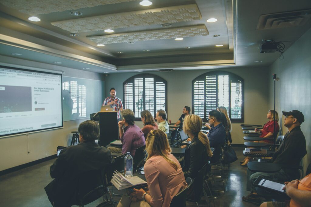 A presenter at the front of a classroom speaking to seated students beside a projected screen