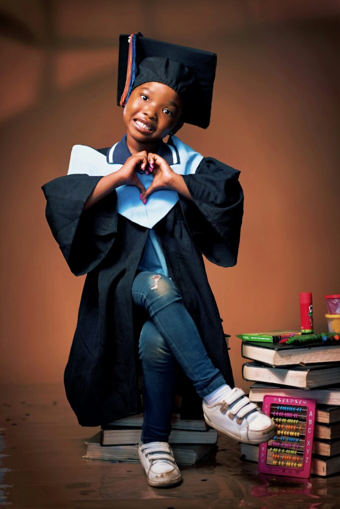 A young graduate in a cap and gown making a heart shape with their hands beside a stack of books