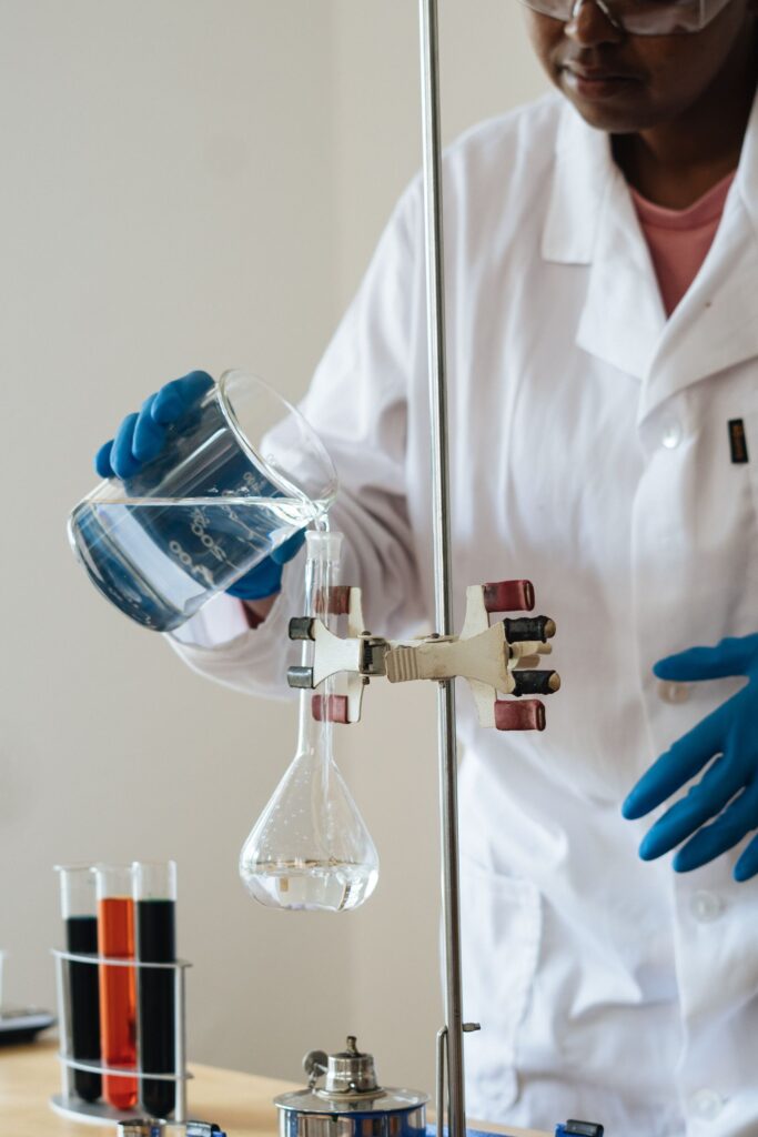 Gloved student in a lab coat pouring liquid from a beaker into a flask on a clamp stand