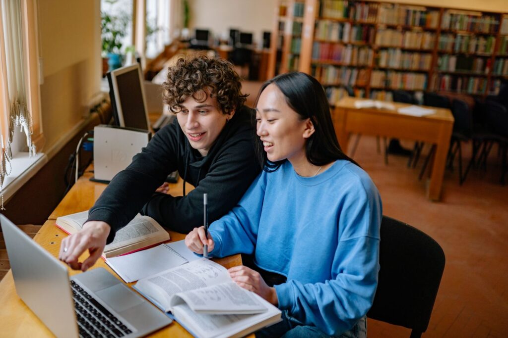 Two students studying together with an open book and laptop in a library