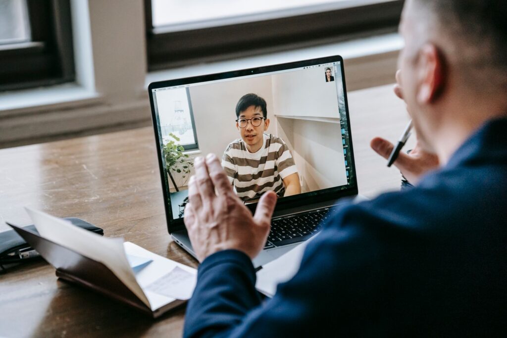 Tutor on a laptop video call guiding a student, with a notebook and pen on the desk