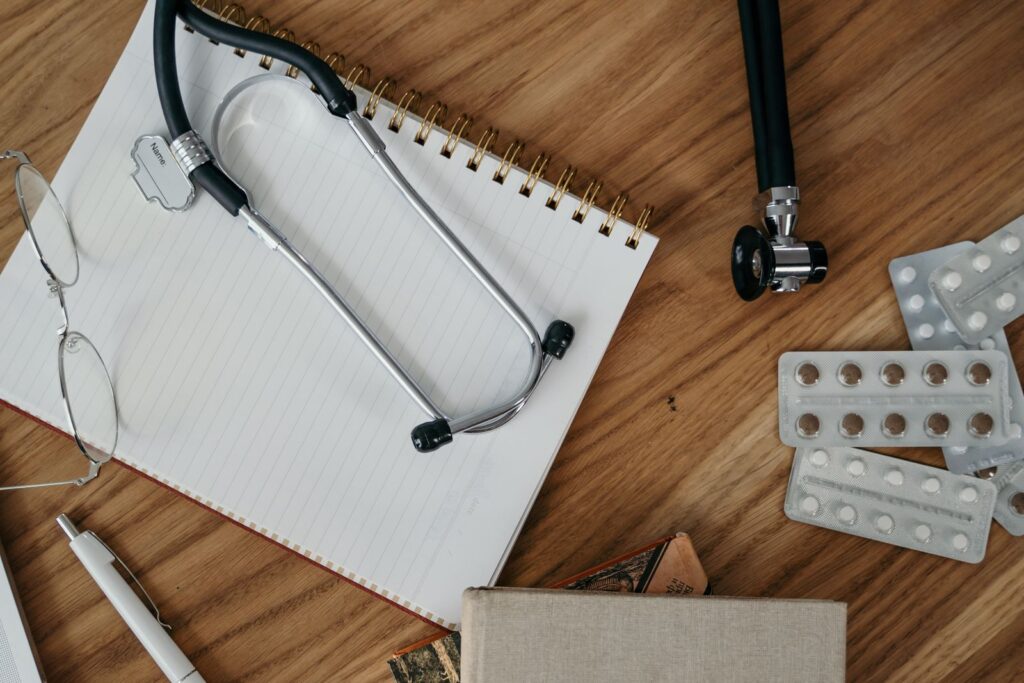 Stethoscope, glasses and a spiral notebook on a wooden desk beside blister packs of tablets